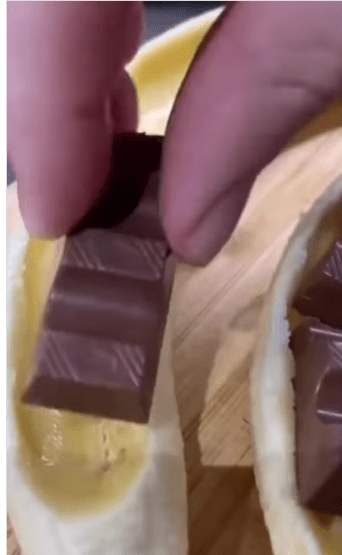 A person is inserting a piece of chocolate into a slit in a banana.